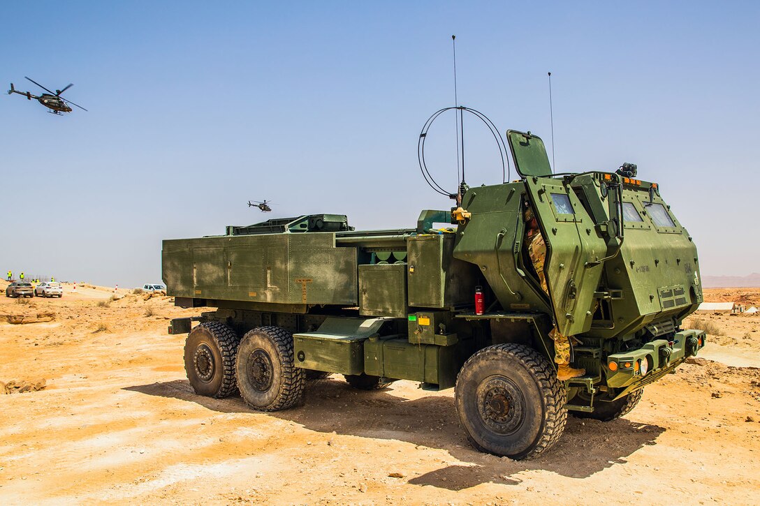 A soldier climbs into a military vehicle in a desert-like area as two helicopters fly nearby.