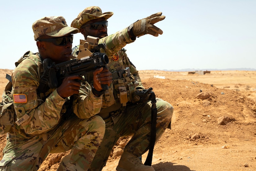 Two soldiers kneel in a barren area, with one pointing and the other aiming a weapon in the same direction.