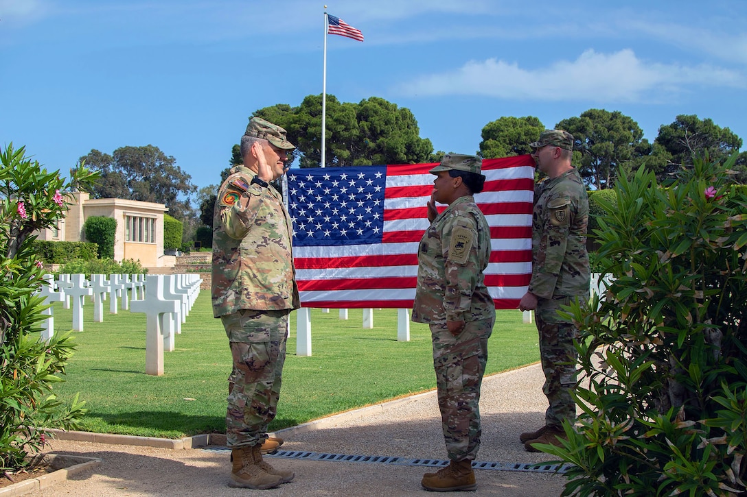 Two soldiers face each other, both holding up their right hands, in front of a U.S. flag others are holding up in a cemetery.