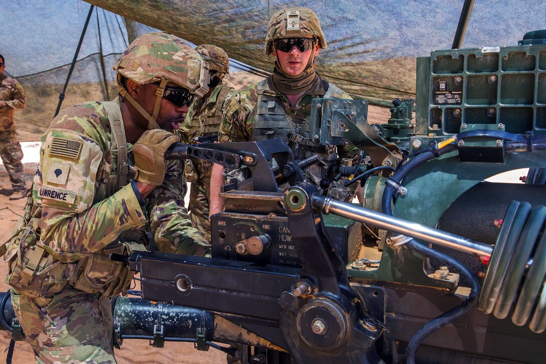 A soldier manipulates a part on a howitzer as others stand nearby under a mesh covering outdoors .