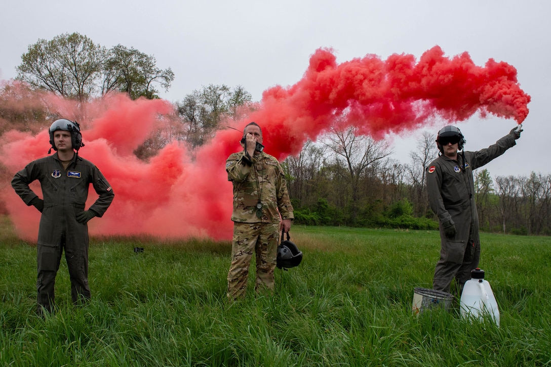 Three airmen stand outside on a cloudy day as one speaks on a walkie-talkie while another launches a red smoke bomb.