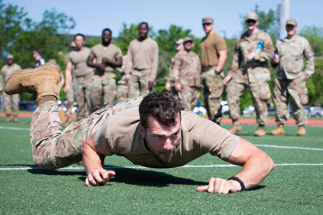 An airman low-crawls during a fitness test on a sunny day as other service members watch in the background.