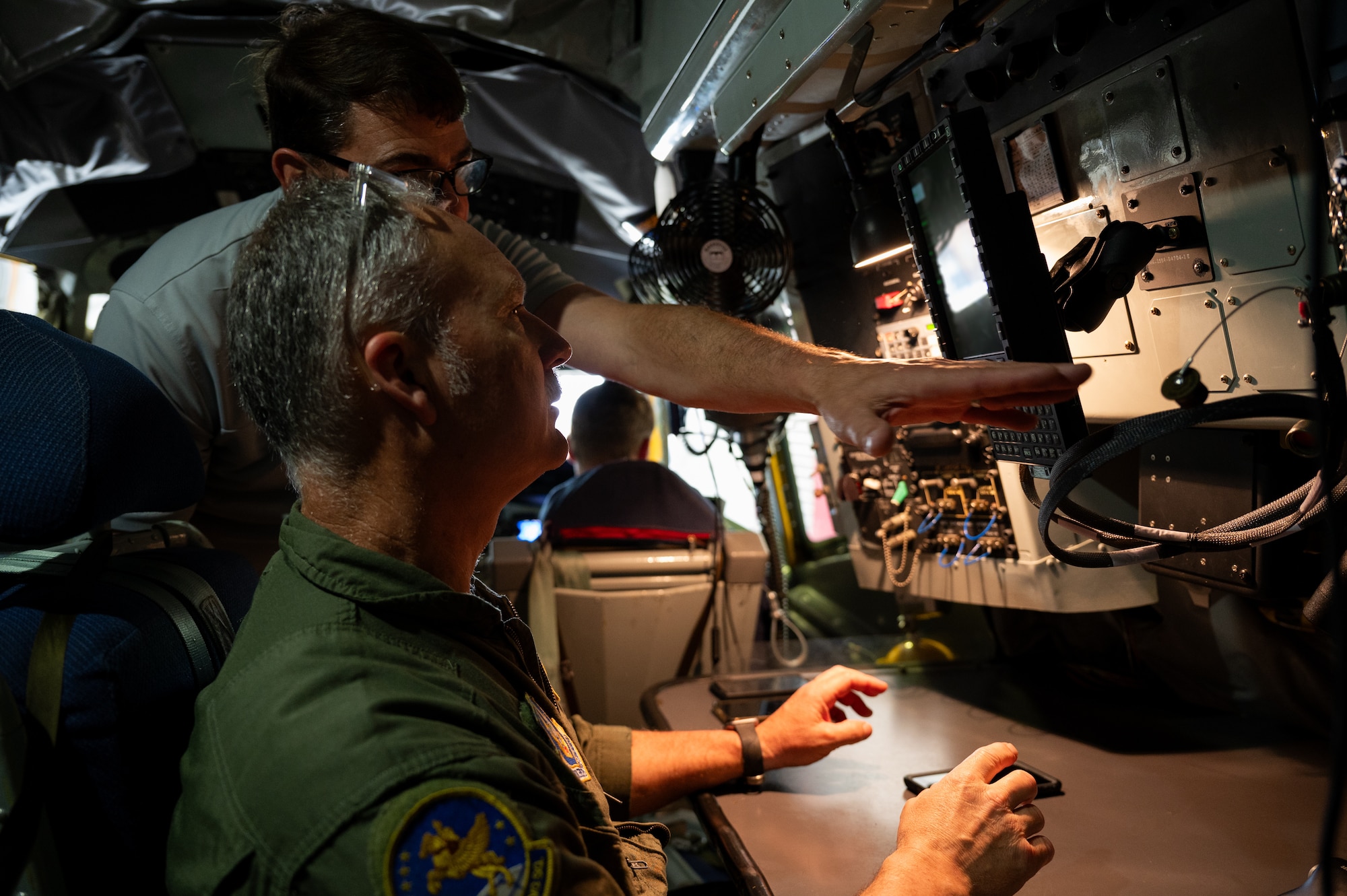 A man in a U.S. Air Force flight uniform sits and looks at a screen that another man, in civilian clothes, is pointing to.