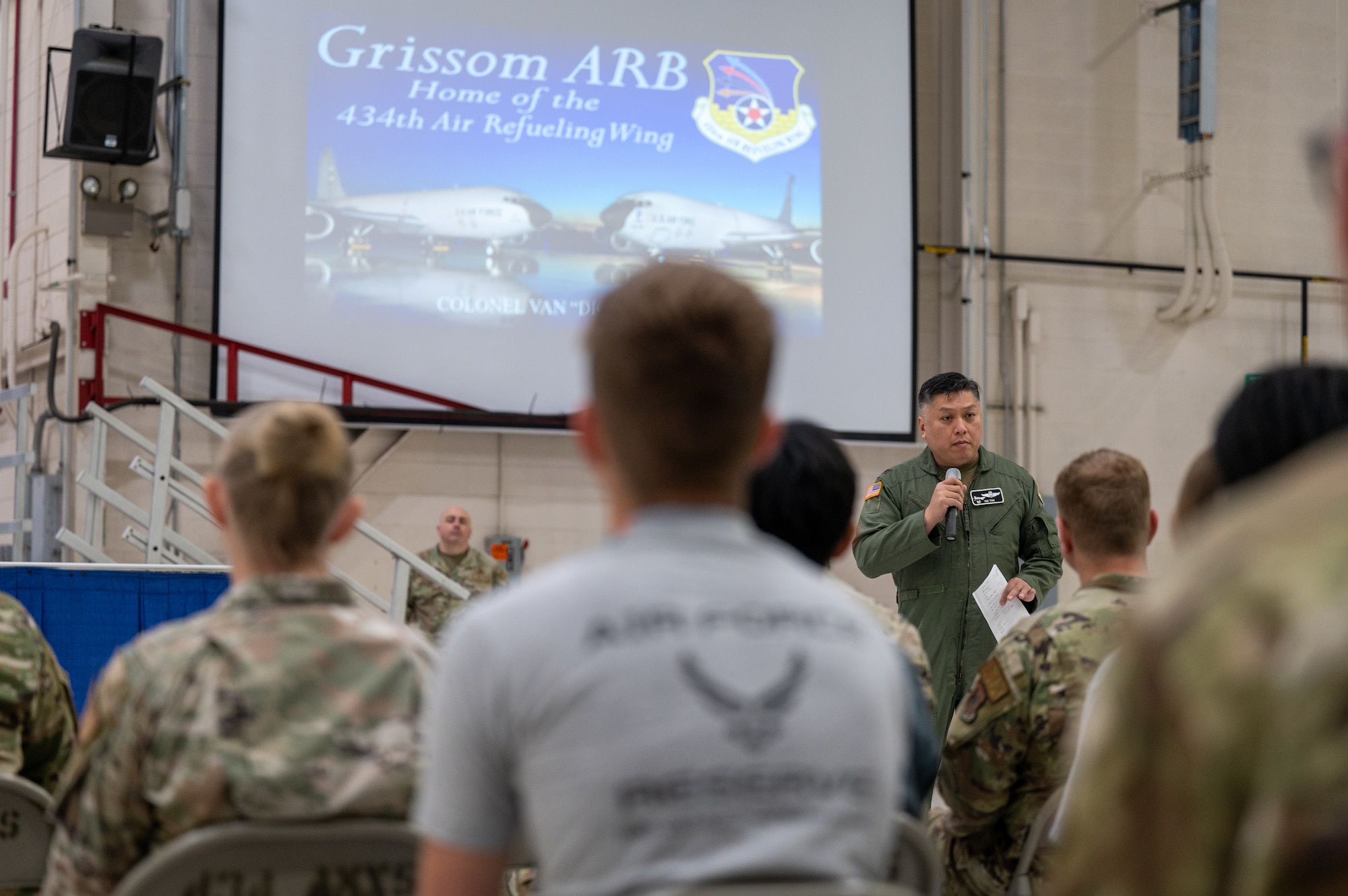 A man in a U.S. Air Force flight suit speaks into a microphone in front of a crowd. The crowd appears to be comprised of those in Operational Camouflage Pattern uniforms as well as some in t-shirts that read "AIR FORCE RESERVE" on the back.