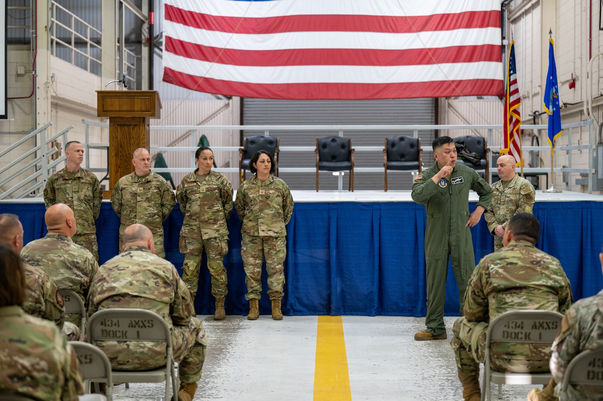 A man in a U.S. Air Force flight suit speaks to a crowd. Beside him, four men and women in Operational Camouflage Pattern uniforms standing in a line, facing the camera. In the foreground, more uniformed servicemembers sitting and listening.