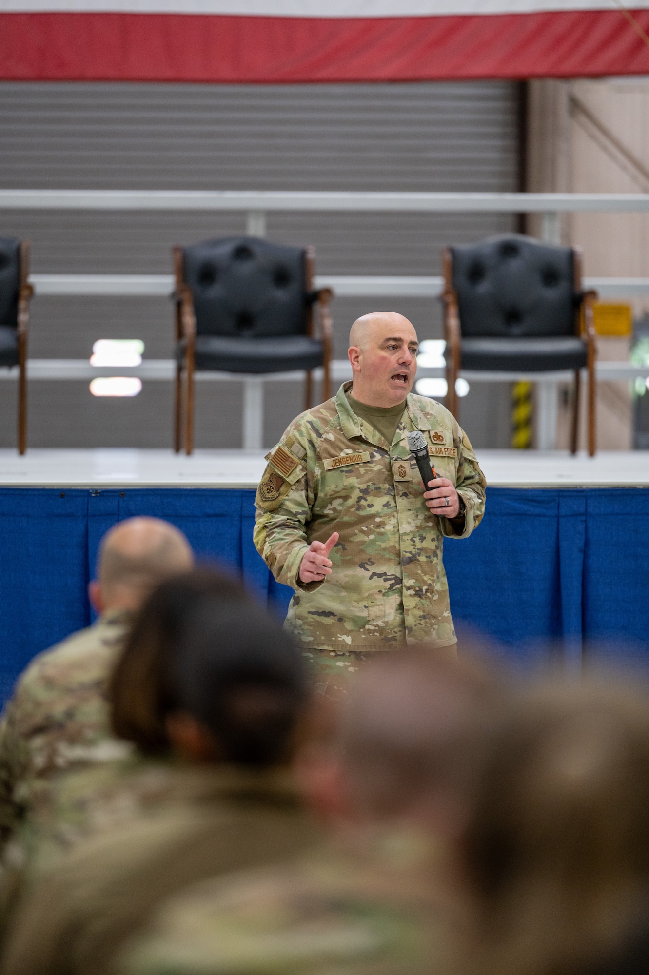 A man in a U.S. Air Force Operational Camouflage Pattern uniform speaks into a microphone. In the foreground, military members sit in the crowd. Behind the speaker, vacant chairs on a stage.