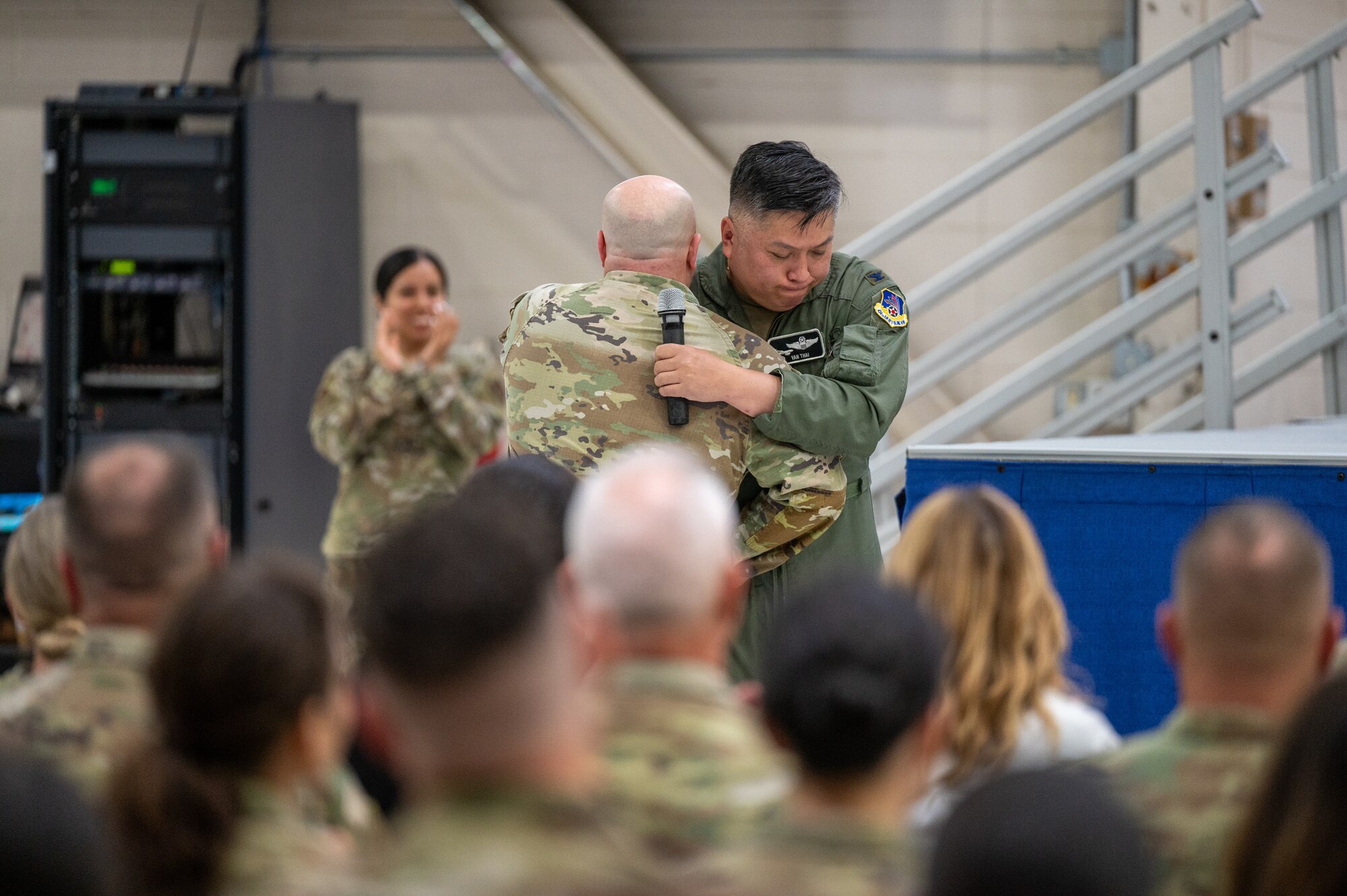 Two men in U.S. Air Force uniforms, one a flight suit and the other a Operational Camouflage Pattern uniform, hug each other. The one in a flight suit is holding a microphone upright.