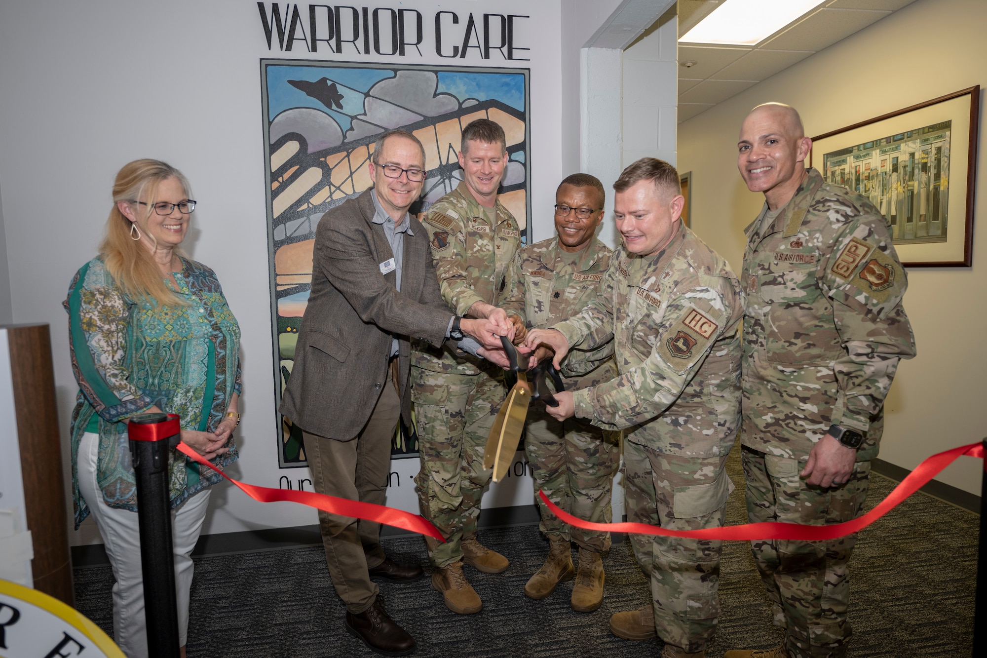 Airmen and two civilians use giant scissors to cut the ribbon in front of the mural.