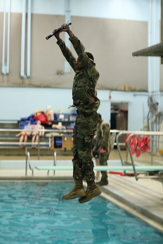 A soldier holding a replica rifle jumping into a pool while suspended in midair.