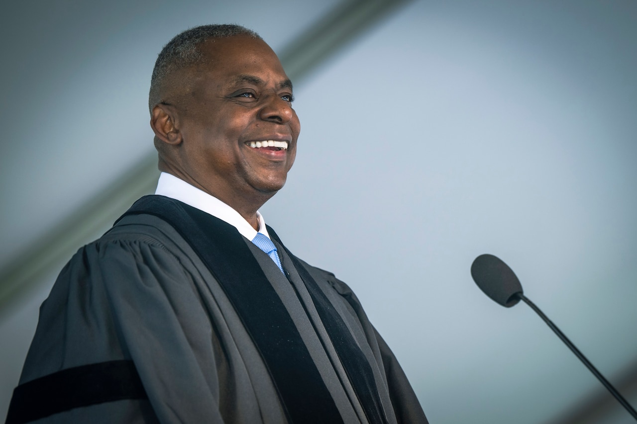 Secretary of Defense Lloyd J. Austin III smiles while wearing a black graduation robe and standing in front of a microphone at a lectern.