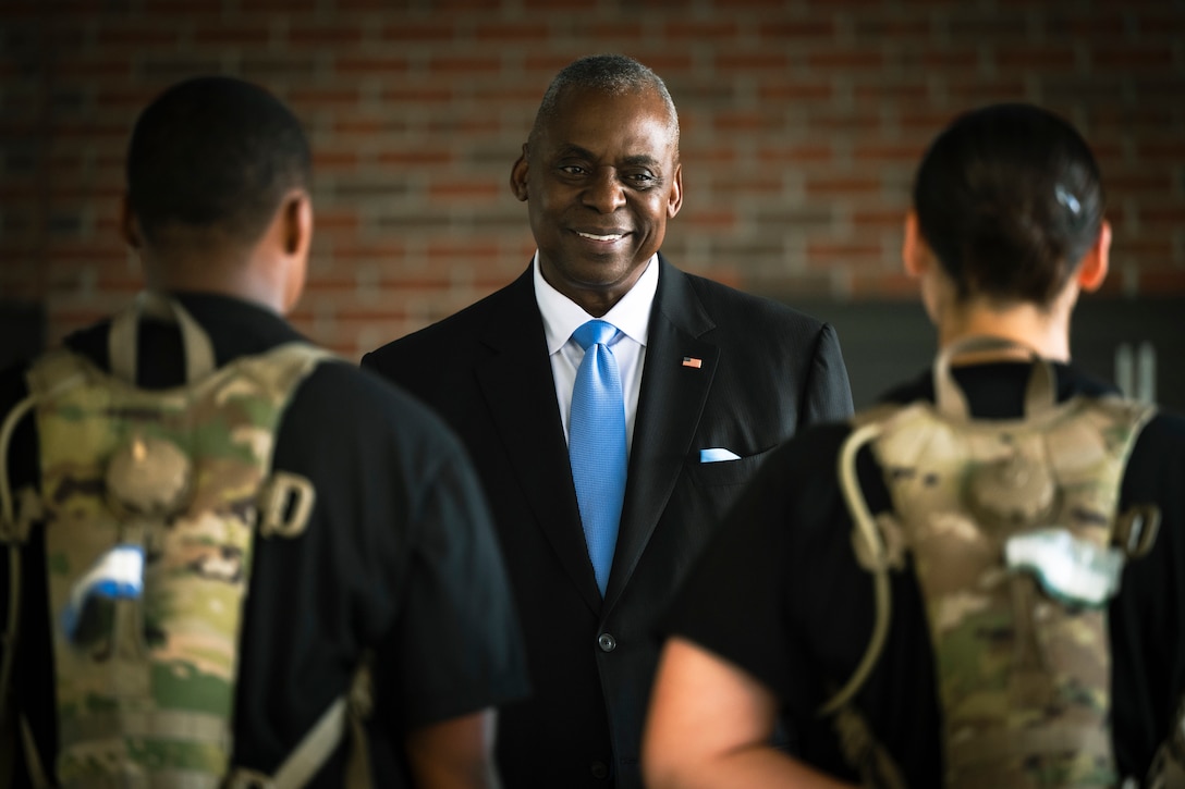 Secretary of Defense Lloyd J. Austin III smiles while speaking to two soldiers wearing camouflage gear on their backs.