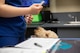 A veterinarian wearing a blue medical outfit is holding a medical device near a light brown dog who is looking up from behind a silver metal operating table.