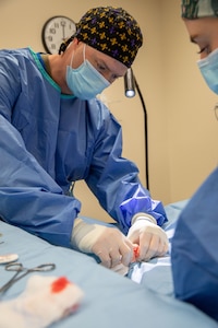 A veterinarian in light blue surgical gown with multicolored cap, white surgical gloves and head-mounted flashlight has his hands on a dog on an operating table covered in light blue cloth.