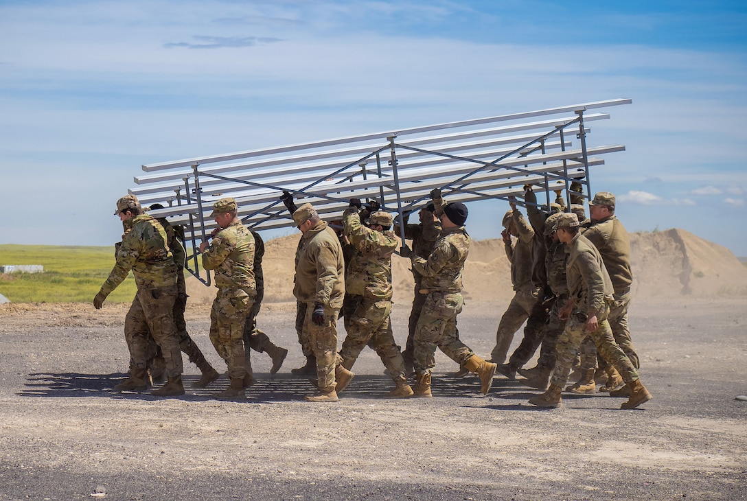 About 15 soldiers carry a section of metal bleachers across a gravel expanse.