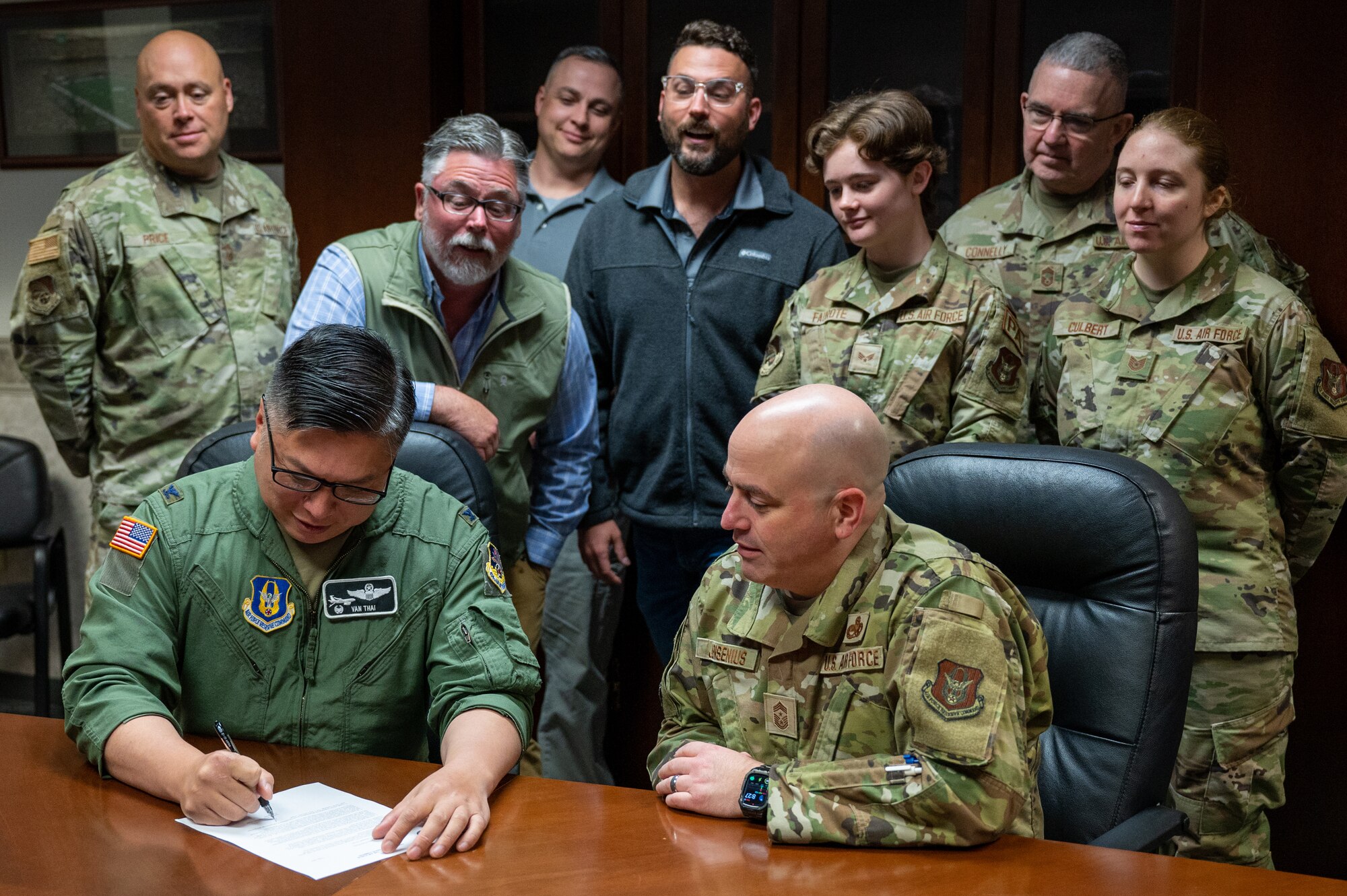 A man in a U.S. Air Force flight uniform signs a memorandum as people crowd around him, looking at the paper.