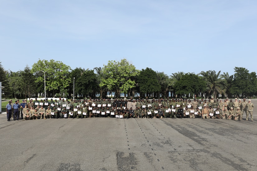 Multinational service members sit and stand for a photo on a paved lot, many displaying certificates.