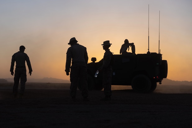 A Marine works on a vehicle as three fellow Marines gather to the left in a desert-like area under a sunlit sky.