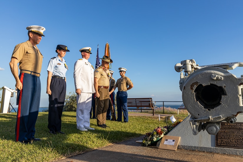 A group of service members stand at attention in front of a memorial during a clear, sunny day.