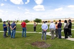 A light skinned woman with blond hair reads a proclamation while standing in a field of grass with newly planted trees. Surrounding her are a large group of people who are listening to her speak.