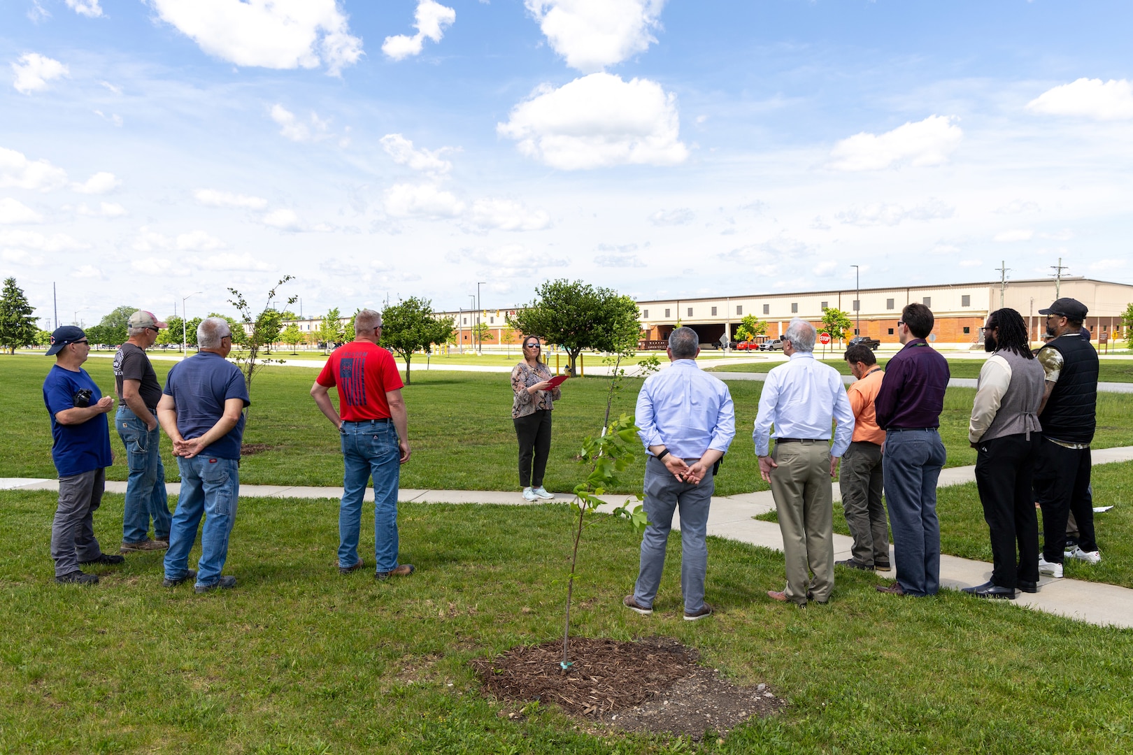 A light skinned woman with blond hair reads a proclamation while standing in a field of grass with newly planted trees. Surrounding her are a large group of people who are listening to her speak.