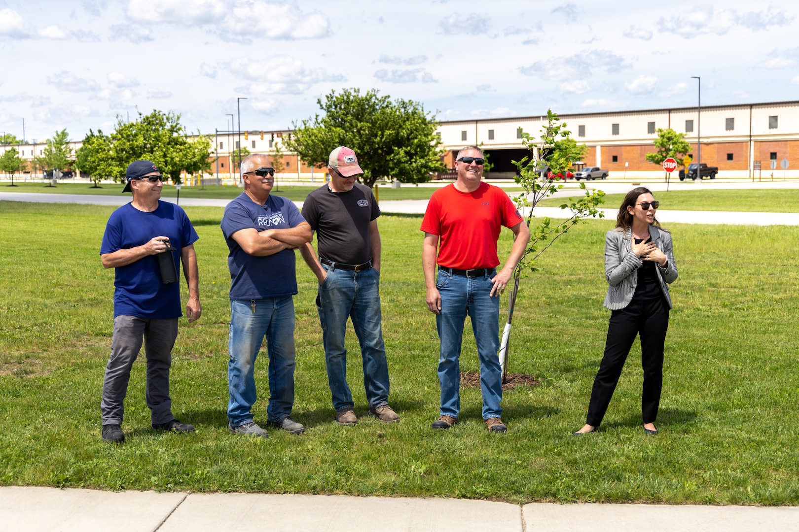 A group of men (four of them) in T-Shirts stand near a few newly planted trees. A light skinned woman in a black and white jacket, black shirt and black pants with sunglasses is speaking.