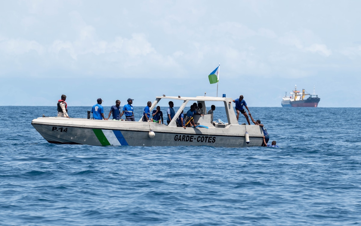 13 people sit on a boat, while two people climb aboard from the back of the vessel.