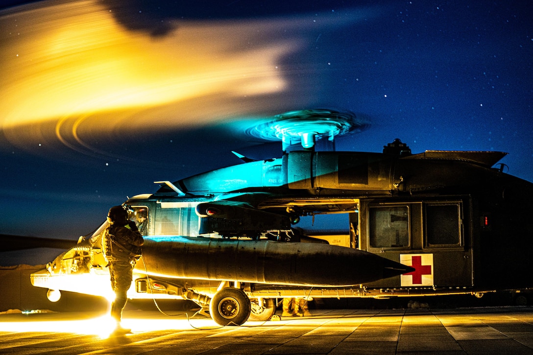 A soldier stands on the tarmac as a military helicopter prepares to fly at night. The photo is illuminated by a yellow light and the propeller is in motion.