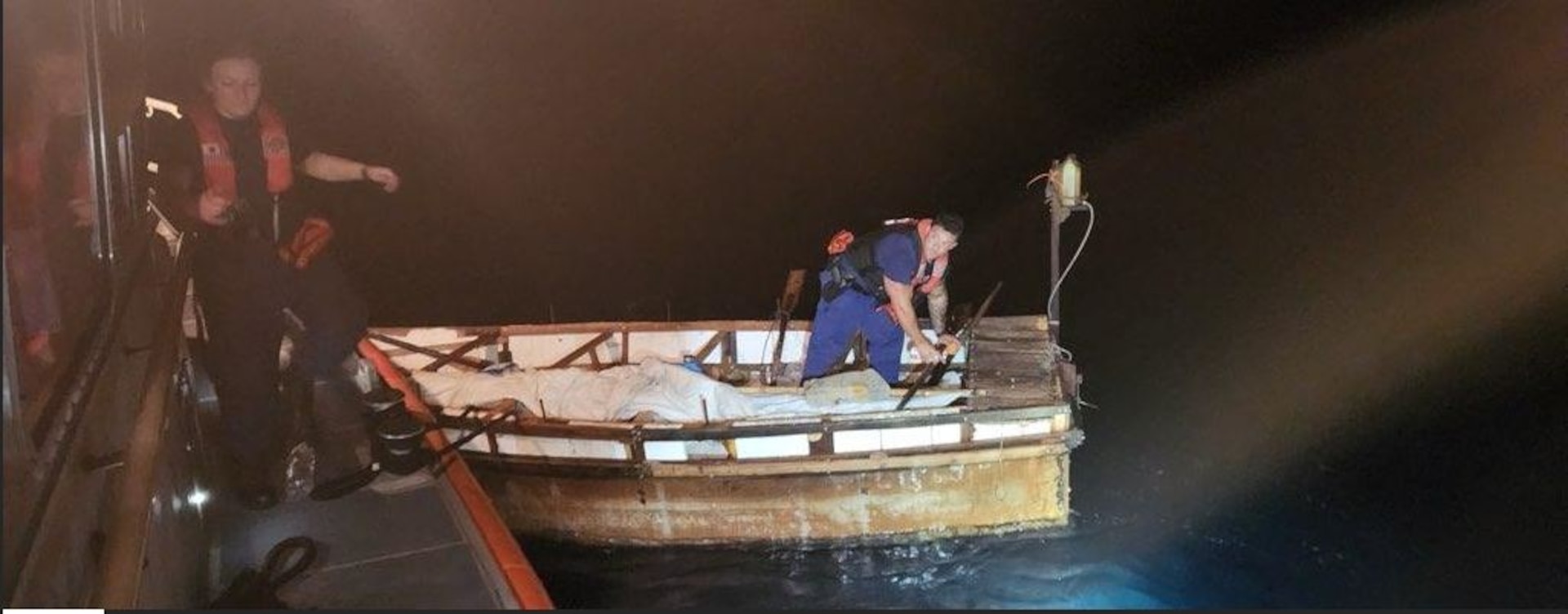 A US Coast Guard boat crew member boards a migrant vessel with spotlight shone on it.
