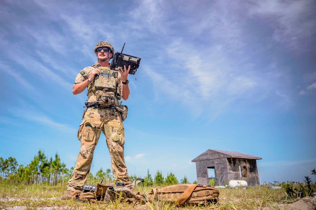 An airman holds a device while a bag and other gear rests at their feet in a field with a small structure and trees in the background.