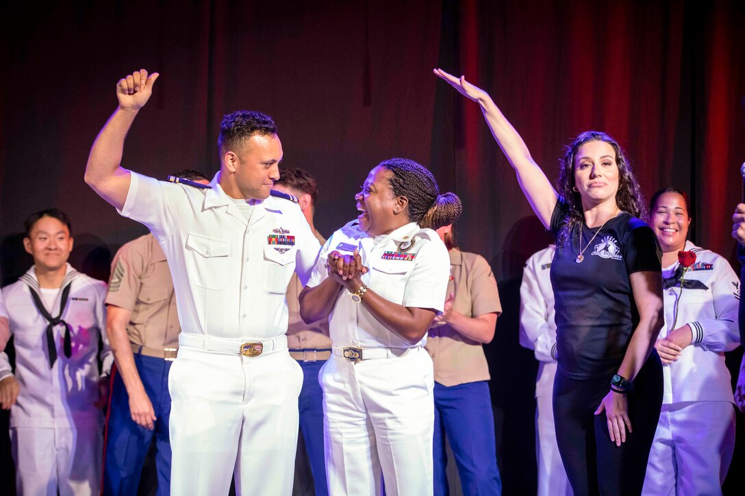 A person holds their hand over two sailors as they excitedly stare at each other while fellow sailors stand behind them in front of a red curtain.