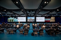 Dozens of people are seated at tables in a large hall facing tall screens during a presentation