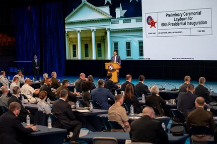 Dozens of people dressed in business attire and uniforms are seated at tables facing large screens while a speaker talks to them. The screens have slides on them, which read "Preliminary Ceremonial Laydown for 60th Presidential Inauguration"