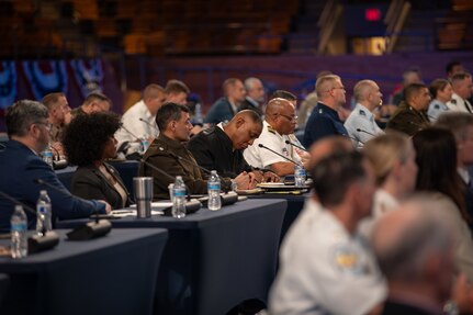 Dozens of people are seated at tables with microphones in front of each person at a meeting.