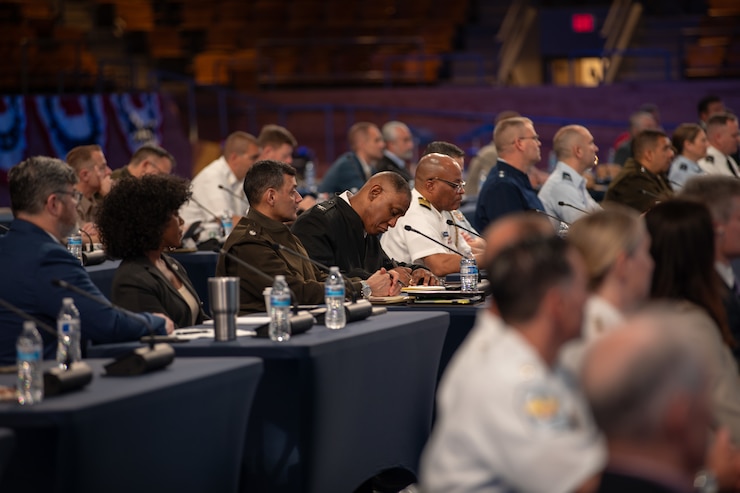 Dozens of people are seated at tables with microphones in front of each person at a meeting.