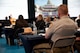 A first responder in dress uniform is seated with a large audience at tables looking at large screens and listening to a speaker.