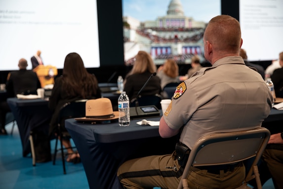 A first responder in dress uniform is seated with a large audience at tables looking at large screens and listening to a speaker.