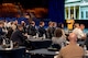 Dozens of people dressed in business attire and military and first responder uniforms are listing to a speaker who is at a wooden podium in front of large screens.