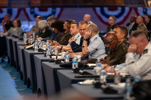 Dozens of people dressed in a mix of military uniforms and business attire are sitting at long tables with microphones in front of each person during a conference.