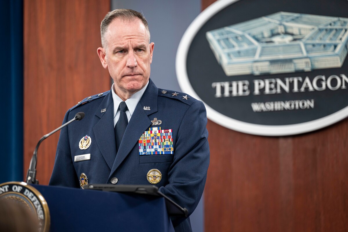 A man in a dress military uniform with multiple award ribbons on his chest and two stars on each shoulder stands at a lectern. Behind him on the wall is a sign with a five-sided building that says, “The Pentagon ... Washington”