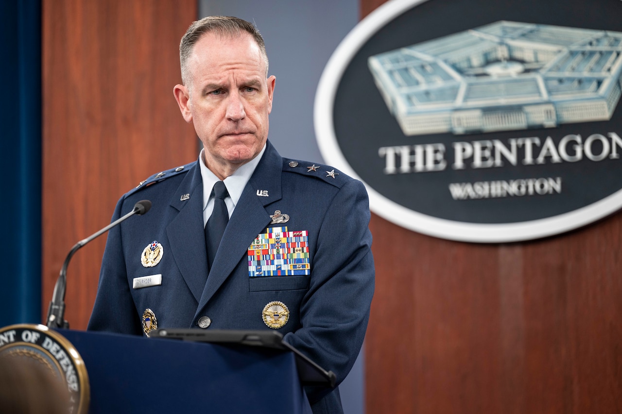 A man in a dress military uniform with multiple award ribbons on his chest and two stars on each shoulder stands at a lectern. Behind him on the wall is a sign with a five-sided building that says, “The Pentagon ... Washington”