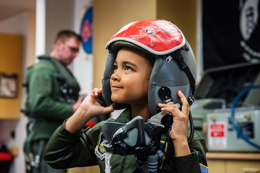 A child, wearing a flight suit, tries on a fighter pilot’s helmet during a visit to a military base.