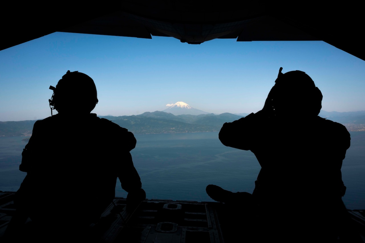 Two airmen are silhouetted against the sky as they sit at the back of an aircraft.