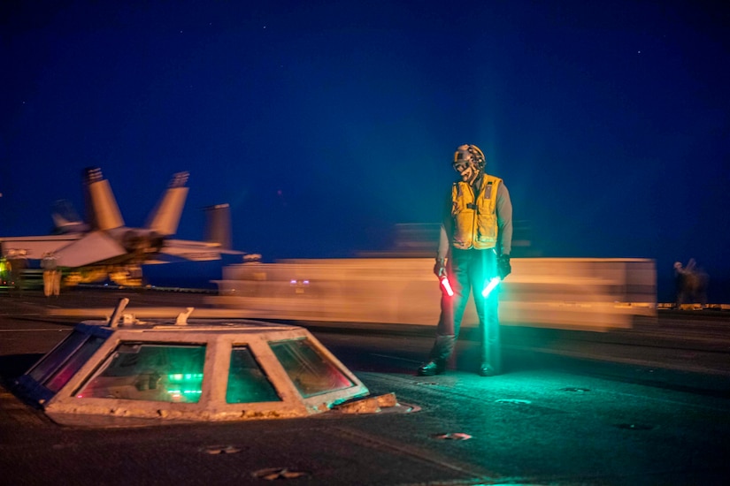 A sailor on a flight deck at night directs a jet.