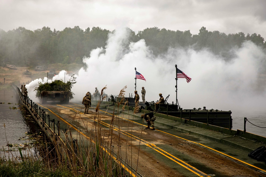 Soldiers work on a bridge displaying two American flags as a military vehicle passes by and smoke arises from a body of water.