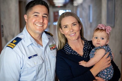 Male service member in uniform smiles next woman smiling and holding baby in her arms.