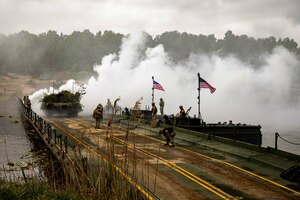 Soldiers work on a bridge displaying two American flags as a military vehicle passes by and smoke arises from a body of water.