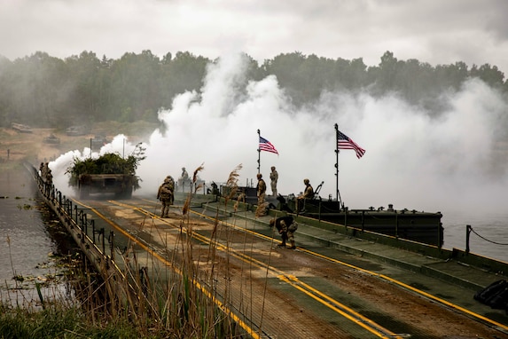 Soldiers work on a bridge displaying two American flags as a military vehicle passes by and smoke arises from a body of water.