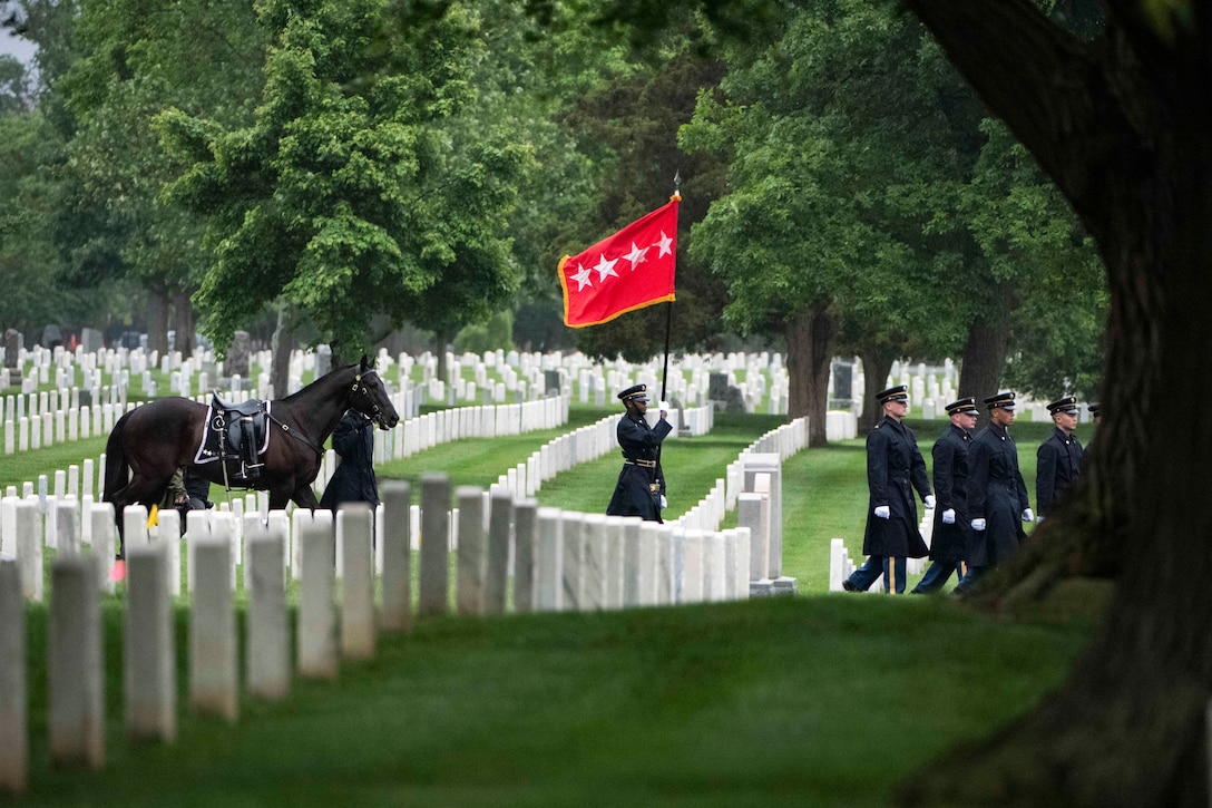 Soldiers in formal uniforms march in formation through a cemetery with one carrying a flag. They’re followed by a horse carrying boots facing backwards in the stirrups.