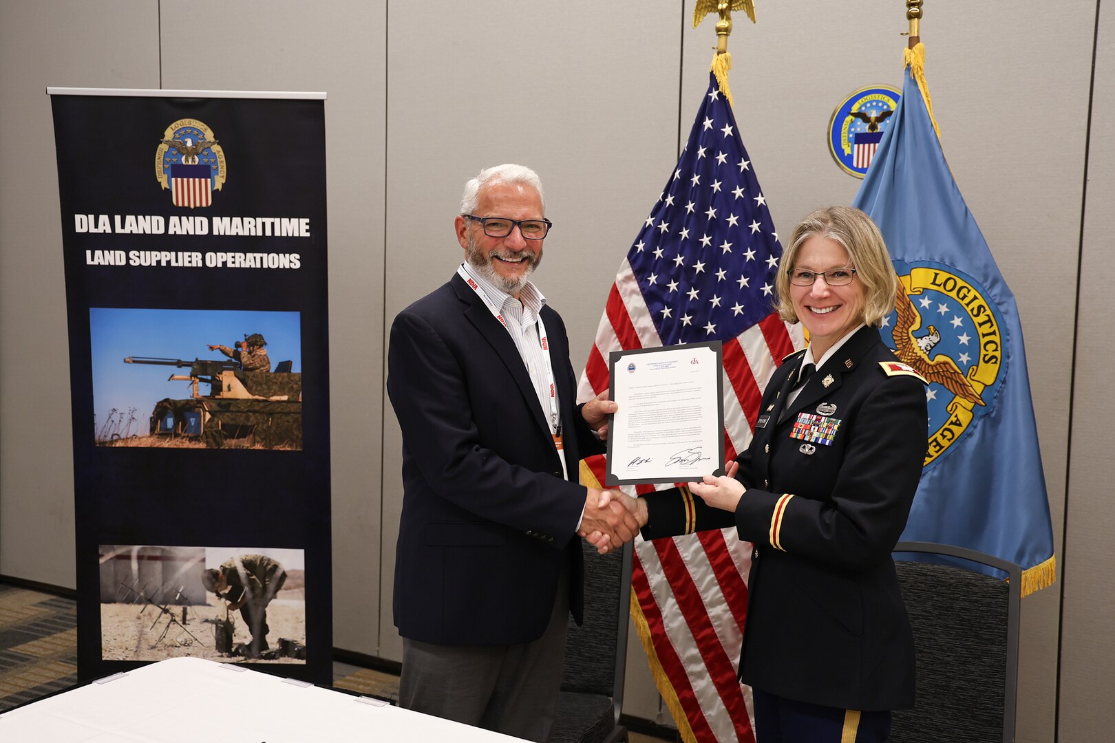 A light skinned woman with blond hair in a dark Army uniform sits and smiles and holds a document with a man in a dark blue suit, also light skinned, also smiling with white hair.