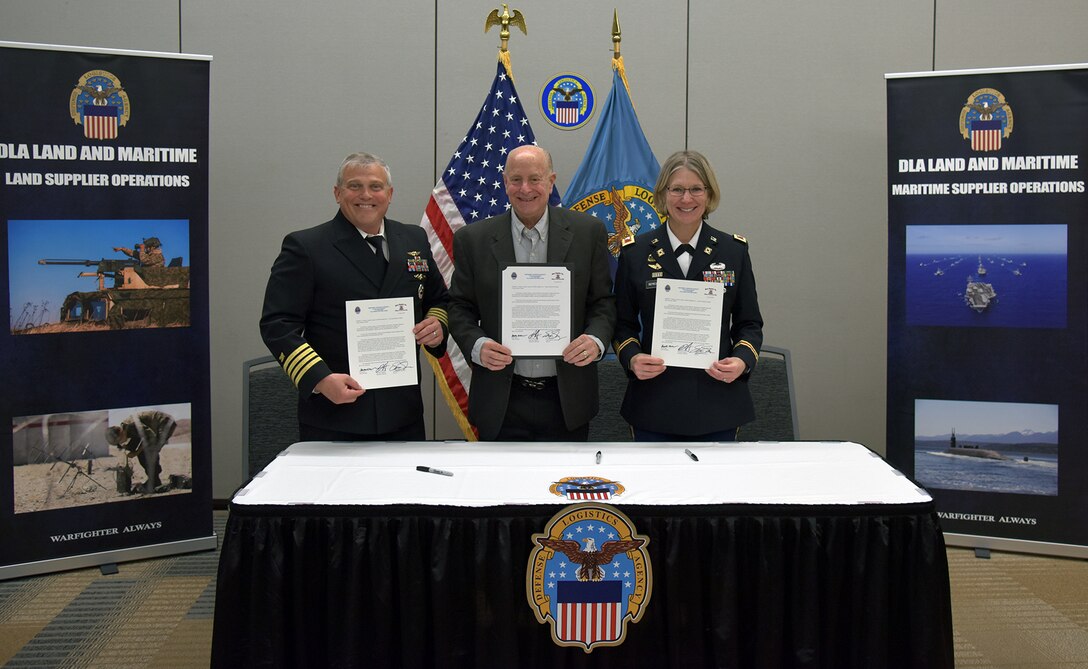 A light skinned woman with blond hair in a dark Army uniform stands and smiles in front of a table with a white tablecloth and a dark skirt with a DLA logo on it with a man in a dark grey suit, also light skinned, also smiling with grey hair and another man in a dark Navy uniform who is light skinned and has grey hair.